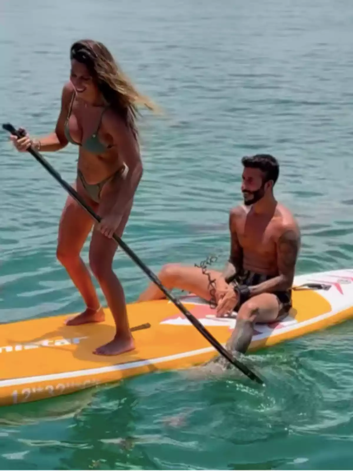 Una mujer y un hombre disfrutan sobre una tabla de paddle surf en el mar, ella de pie remando y él sentado sonriendo Una mujer y un hombre disfrutan sobre una tabla de paddle surf en el mar, ella de pie remando y él sentado sonriendo