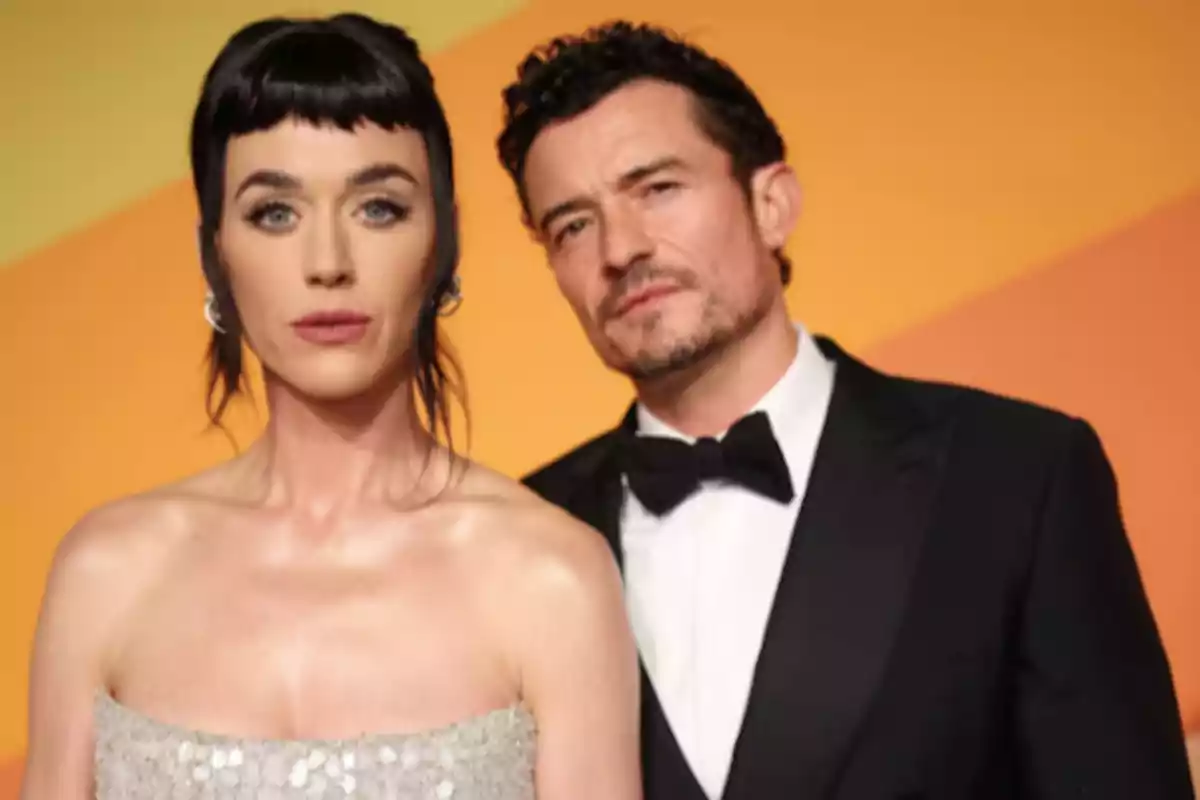 A woman in a silver dress and a man in a tuxedo pose together in front of an orange background.