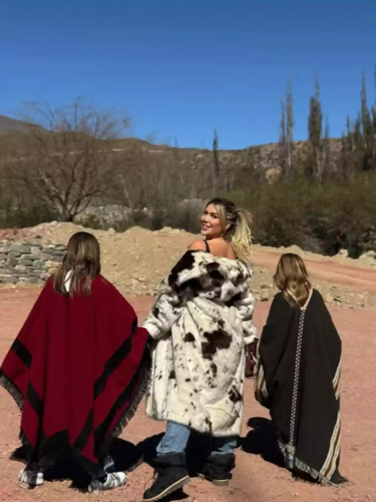 Three people walk away in a dry, mountainous landscape; one of them is wearing a fur coat and the other two are wearing ponchos, while the sky is clear. Three people walk away in a dry, mountainous landscape; one of them is wearing a fur coat and the other two are wearing ponchos, while the sky is clear.