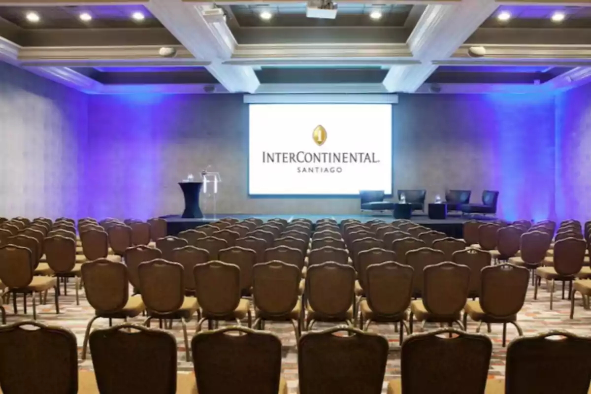 Conference room with rows of empty chairs facing a stage with a large screen and blue lights on the walls Conference room with rows of empty chairs facing a stage with a large screen and blue lights on the walls