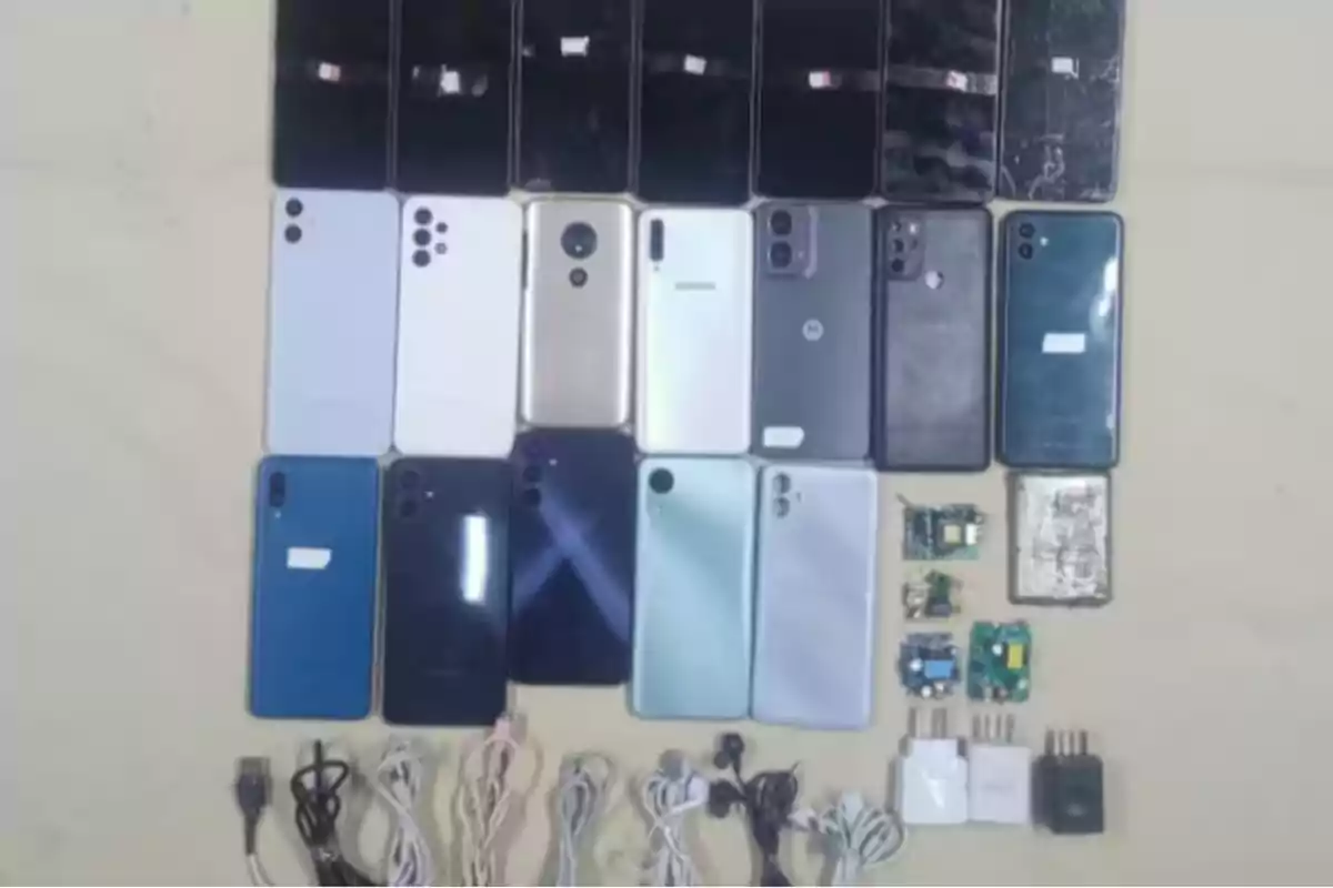 Several mobile phones of different models and colors, accompanied by cables, headphones, chargers, and some electronic boards, all organized on a light-colored surface.