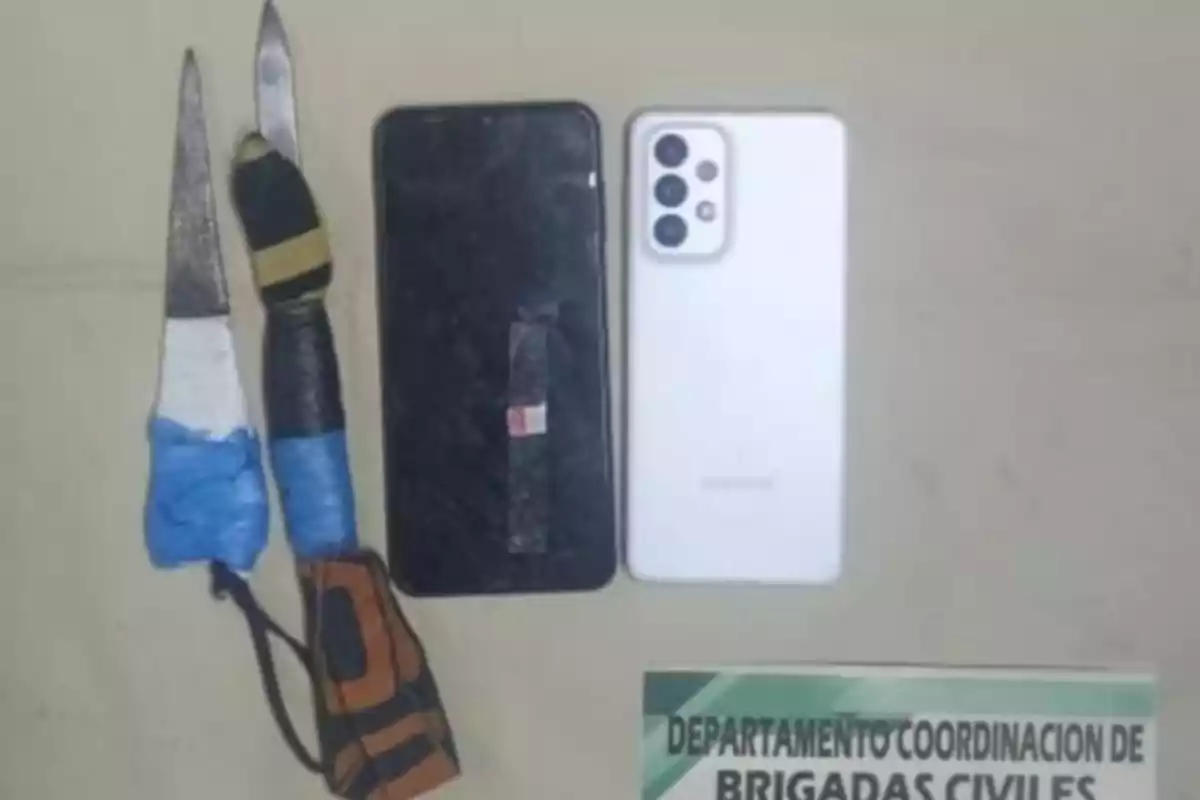 Two homemade knives and two cell phones on a light-colored surface next to a partial sign from the civil brigades department. Two homemade knives and two cell phones on a light-colored surface next to a partial sign from the civil brigades department.