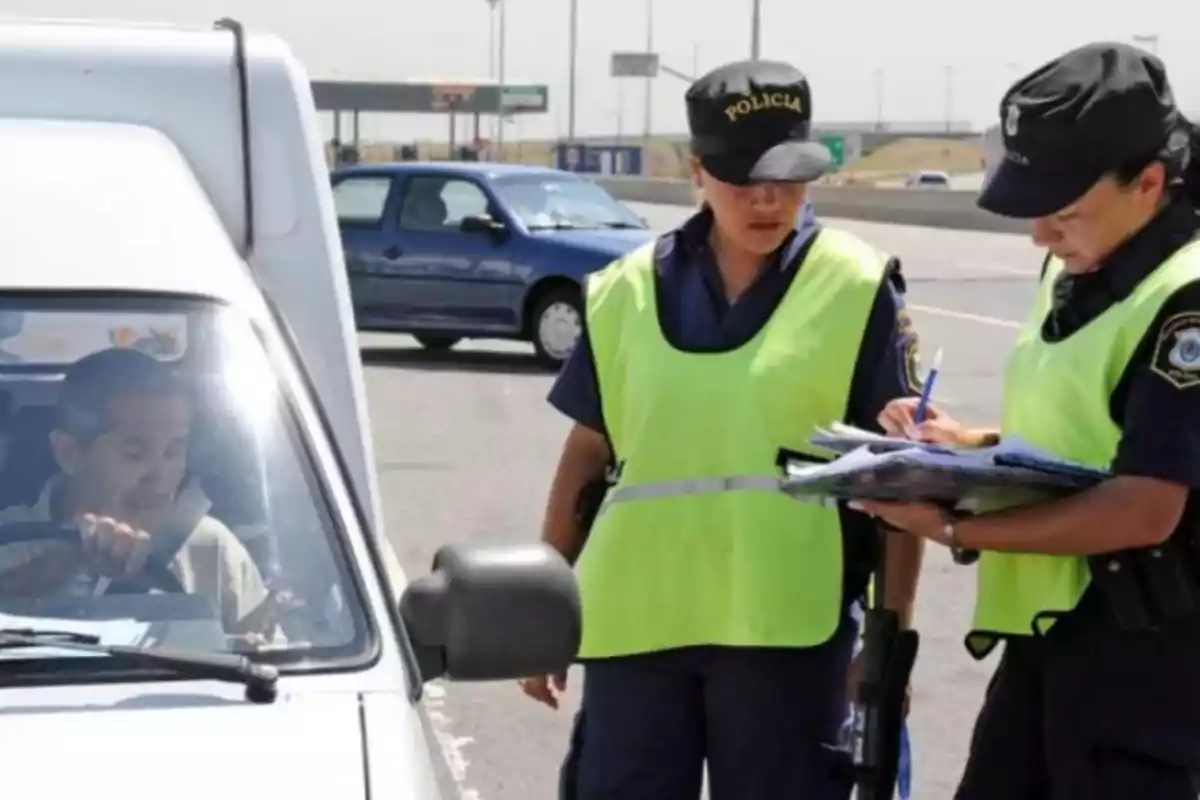 Two traffic police officers wearing reflective vests check documents and take notes next to a car stopped at a roadside checkpoint. Two traffic police officers wearing reflective vests check documents and take notes next to a car stopped at a roadside checkpoint.