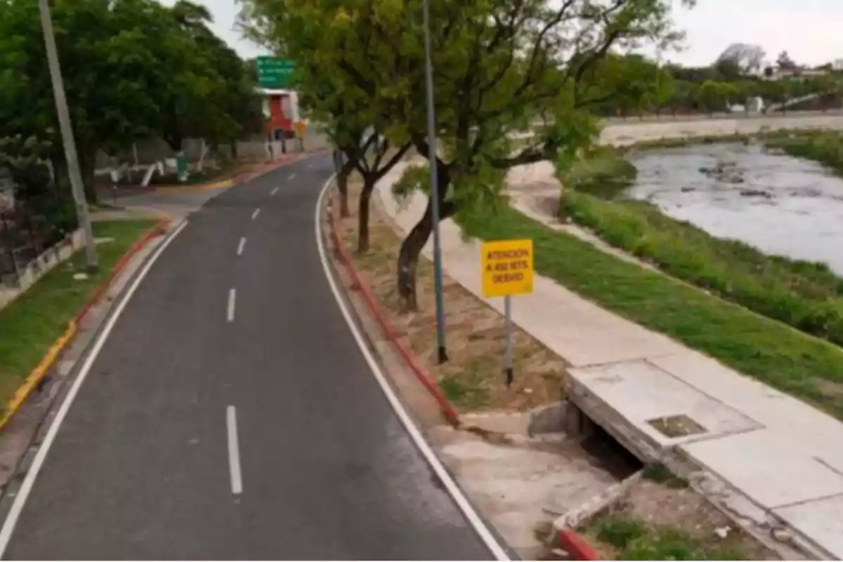 Carretera asfaltada con señal amarilla de advertencia junto a un río y árboles Carretera asfaltada con señal amarilla de advertencia junto a un río y árboles
