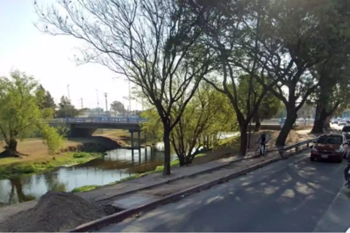 Una calle junto a un río con árboles a los lados, un puente al fondo y un ciclista circulando por la banqueta Una calle junto a un río con árboles a los lados, un puente al fondo y un ciclista circulando por la banqueta