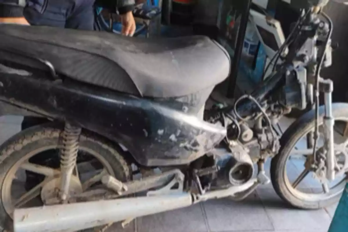 Motocicleta negra en mal estado con partes faltantes y sucia, estacionada en un taller o local con una persona de pie al fondo. Motocicleta negra en mal estado con partes faltantes y sucia, estacionada en un taller o local con una persona de pie al fondo.