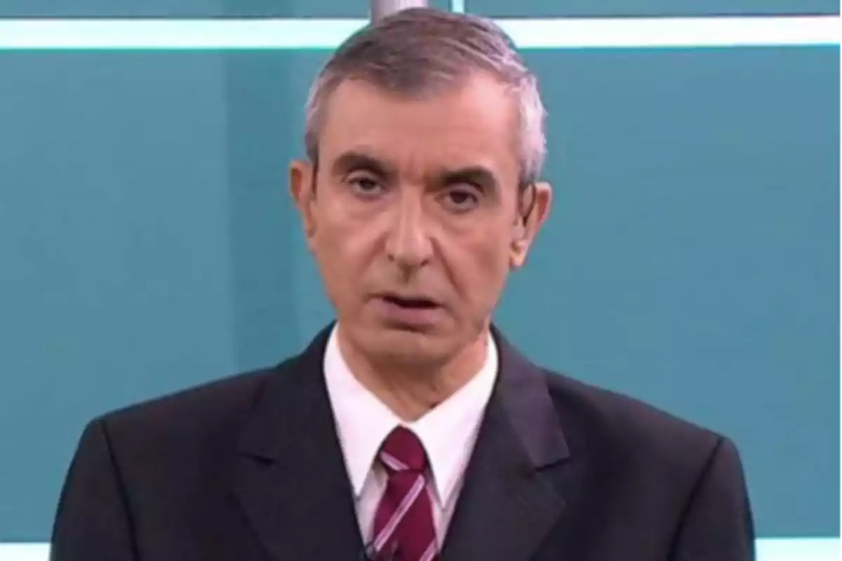 Short gray-haired man wearing a dark suit and red tie in front of a blue background in a television studio