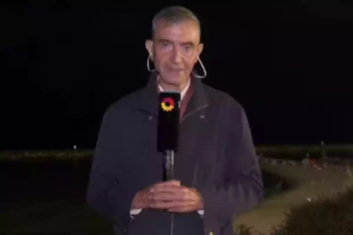 Un hombre de cabello corto sostiene un micrófono con el logo de un canal de televisión mientras está de pie al aire libre durante la noche