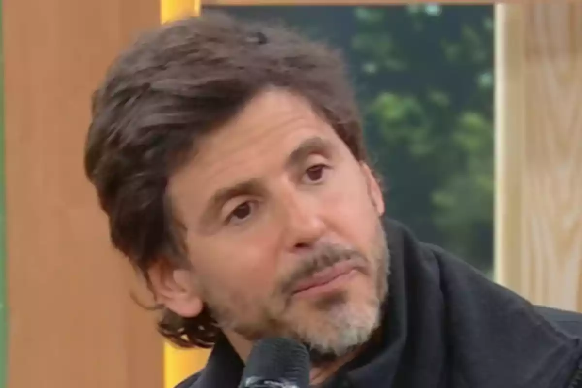 Hombre de cabello castaño y barba corta sosteniendo un micrófono durante una entrevista en un estudio de televisión Hombre de cabello castaño y barba corta sosteniendo un micrófono durante una entrevista en un estudio de televisión
