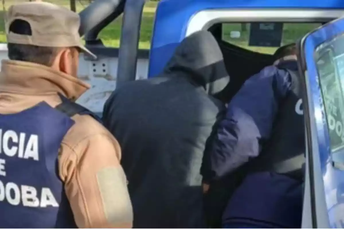 A police officer escorts a hooded person into a patrol car while another officer watches.