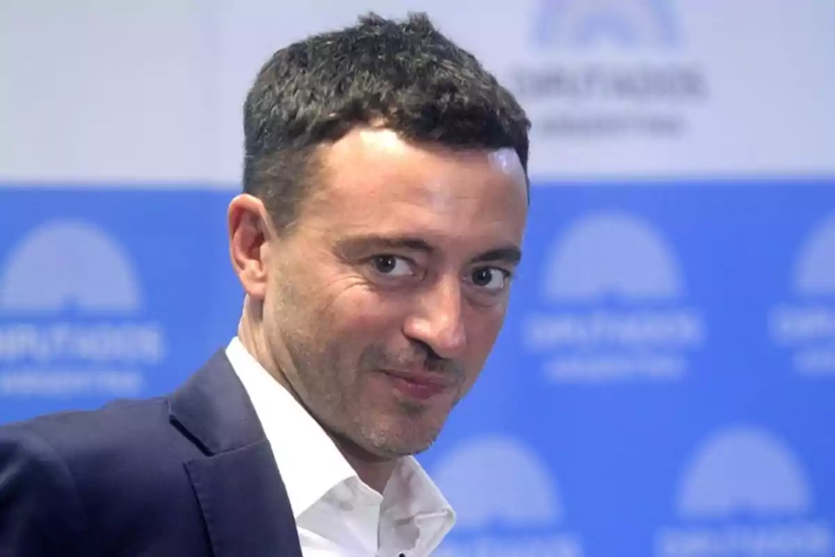 Hombre de cabello corto y oscuro con saco azul y camisa blanca sonriendo levemente frente a un fondo azul con logotipos desenfocados.