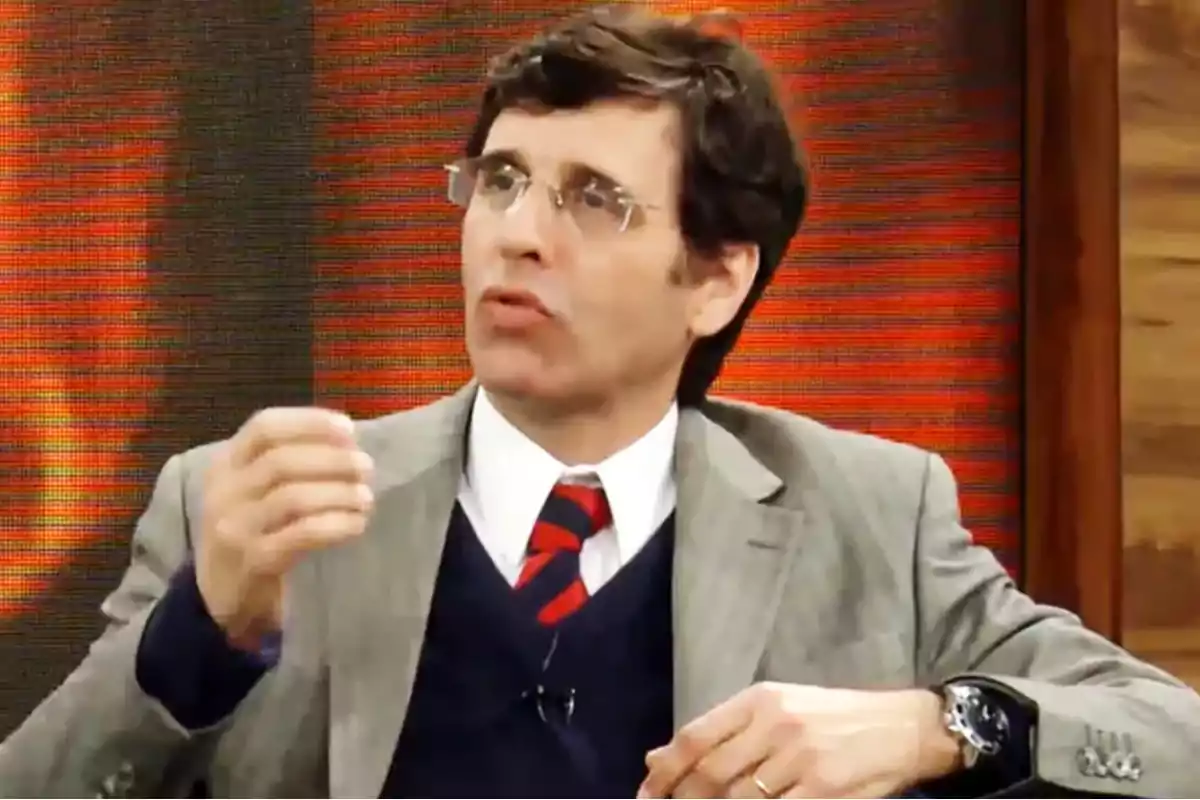 Hombre con gafas y traje gris hablando en un programa de televisión con fondo rojo
