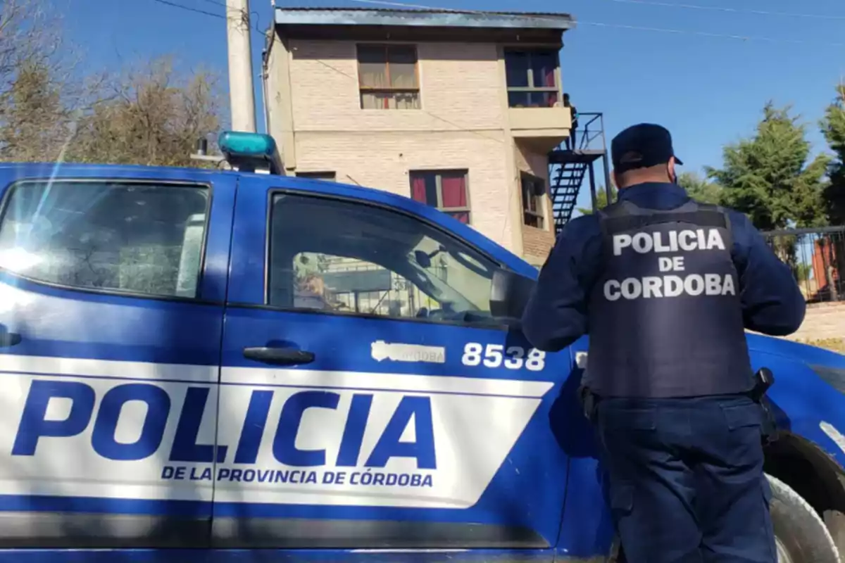 Un policía de Córdoba de espaldas junto a una patrulla azul estacionada frente a un edificio de dos pisos