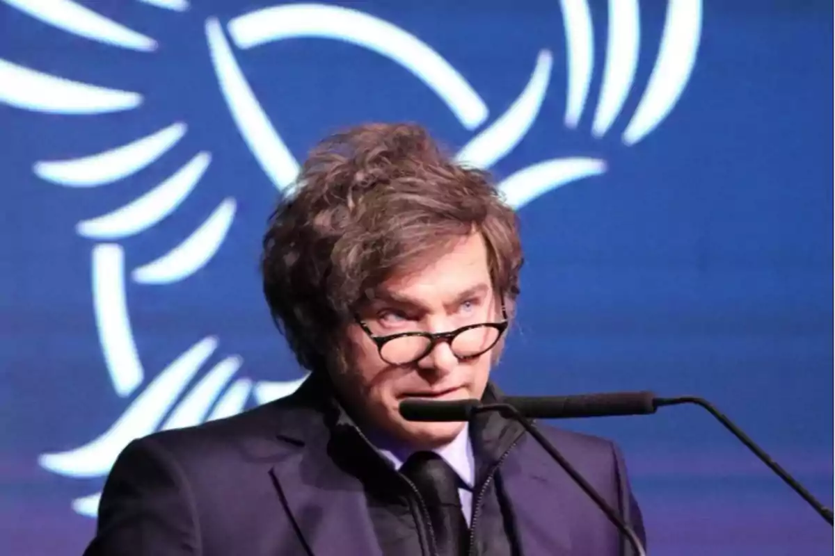 Disheveled-haired man with glasses speaking in front of a microphone with a blue background and abstract white design.