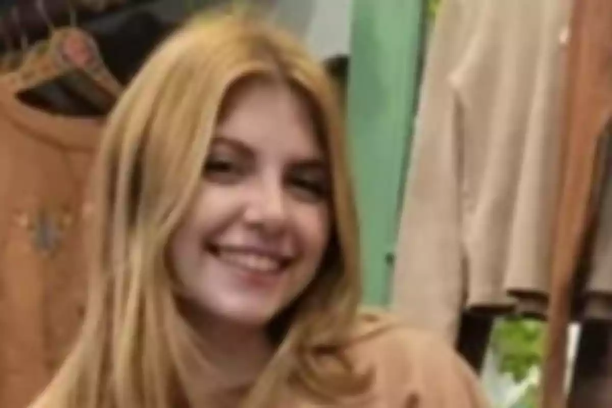 Young blonde-haired person smiling in a clothing store with garments hanging in the background