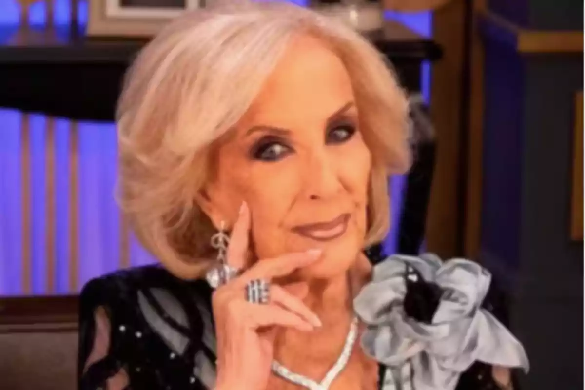 Older woman with short blonde hair, elegantly dressed with jewelry and a gray flower on her shoulder, posing with her hand on her face and a background lit in blue tones