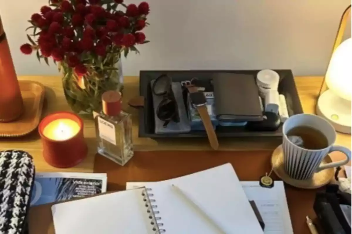 Escritorio organizado con una libreta abierta, una taza de té, una vela encendida, un ramo de flores rojas, una lámpara, perfume, gafas, reloj, cartera y otros objetos personales Escritorio organizado con una libreta abierta, una taza de té, una vela encendida, un ramo de flores rojas, una lámpara, perfume, gafas, reloj, cartera y otros objetos personales