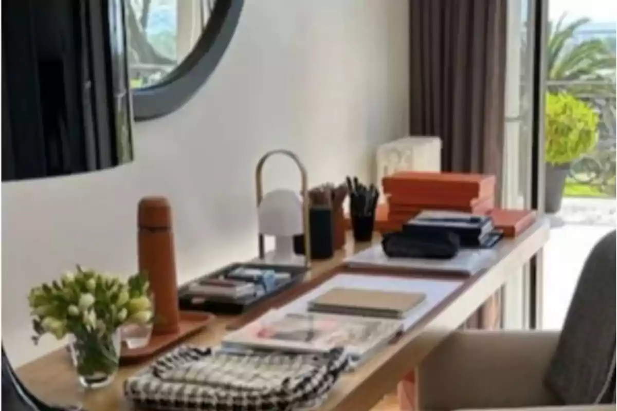 Escritorio de madera con varios objetos organizados como cuadernos, cajas naranjas, una lámpara pequeña, un termo, flores y artículos de papelería junto a una ventana con vista al exterior Escritorio de madera con varios objetos organizados como cuadernos, cajas naranjas, una lámpara pequeña, un termo, flores y artículos de papelería junto a una ventana con vista al exterior