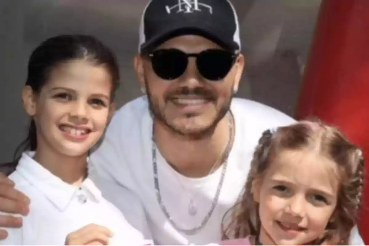 Un hombre con gafas de sol y gorra sonríe junto a dos niñas pequeñas que también están sonriendo Un hombre con gafas de sol y gorra sonríe junto a dos niñas pequeñas que también están sonriendo