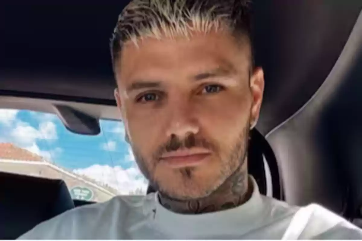 Hombre joven con barba y cabello corto rubio sentado dentro de un automóvil con el cielo azul visible por la ventana trasera