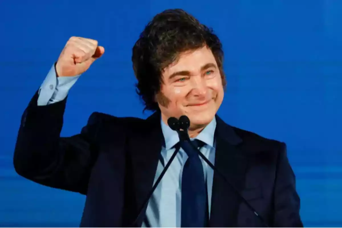 Hombre de cabello oscuro con traje y corbata azul levantando el puño derecho y sonriendo frente a un fondo azul