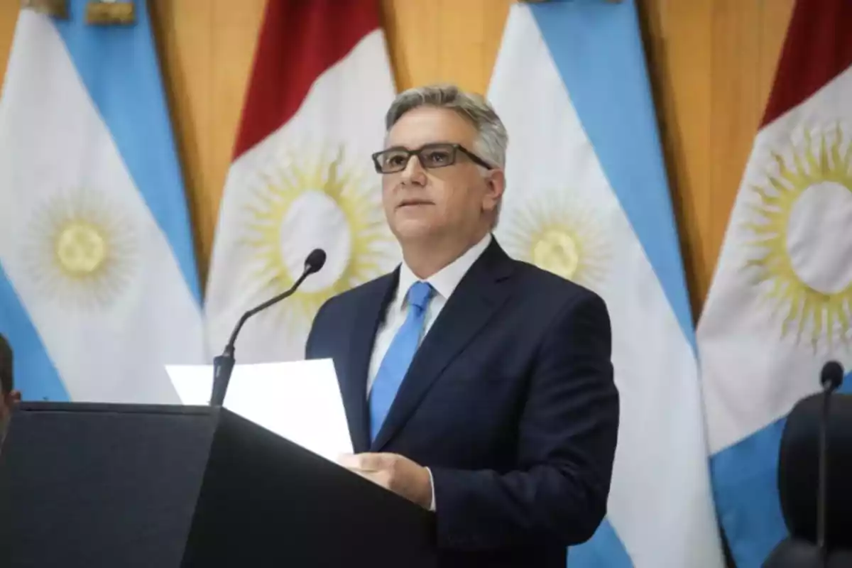 Hombre de traje y corbata azul hablando en un podio con banderas argentinas de fondo