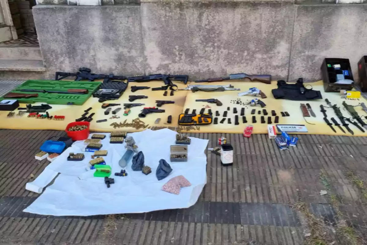 Varias armas de fuego, municiones, cargadores, chaleco antibalas, cuchillos y otros objetos están organizados sobre mantas en el suelo frente a una pared de cemento.