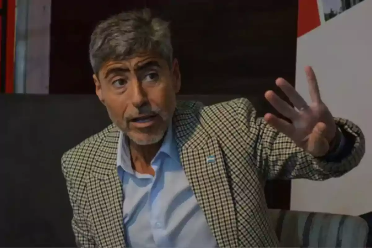 Hombre de cabello canoso y barba corta usando saco a cuadros y camisa celeste gesticulando con la mano derecha mientras habla