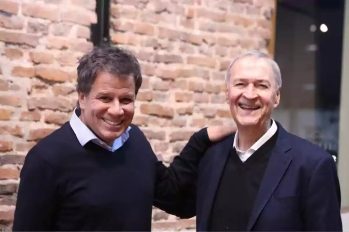 Dos hombres sonrientes posan juntos frente a una pared de ladrillos Dos hombres sonrientes posan juntos frente a una pared de ladrillos