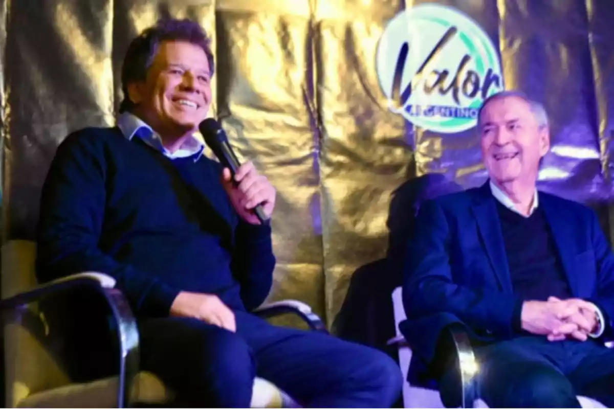 Dos hombres sentados en sillas sonríen durante un evento, uno de ellos sostiene un micrófono y detrás hay un fondo dorado con un logo que dice Valor