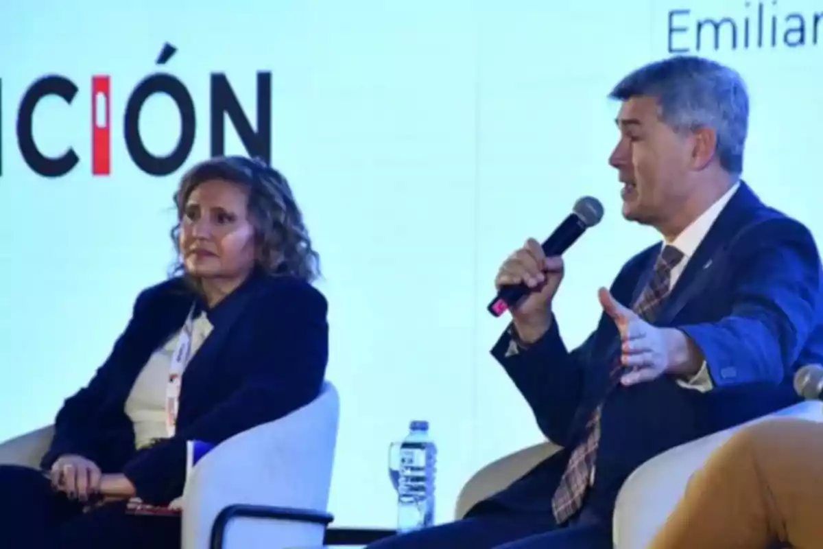 Dos personas sentadas en sillones participan en un panel, una mujer escucha mientras un hombre habla usando un micrófono