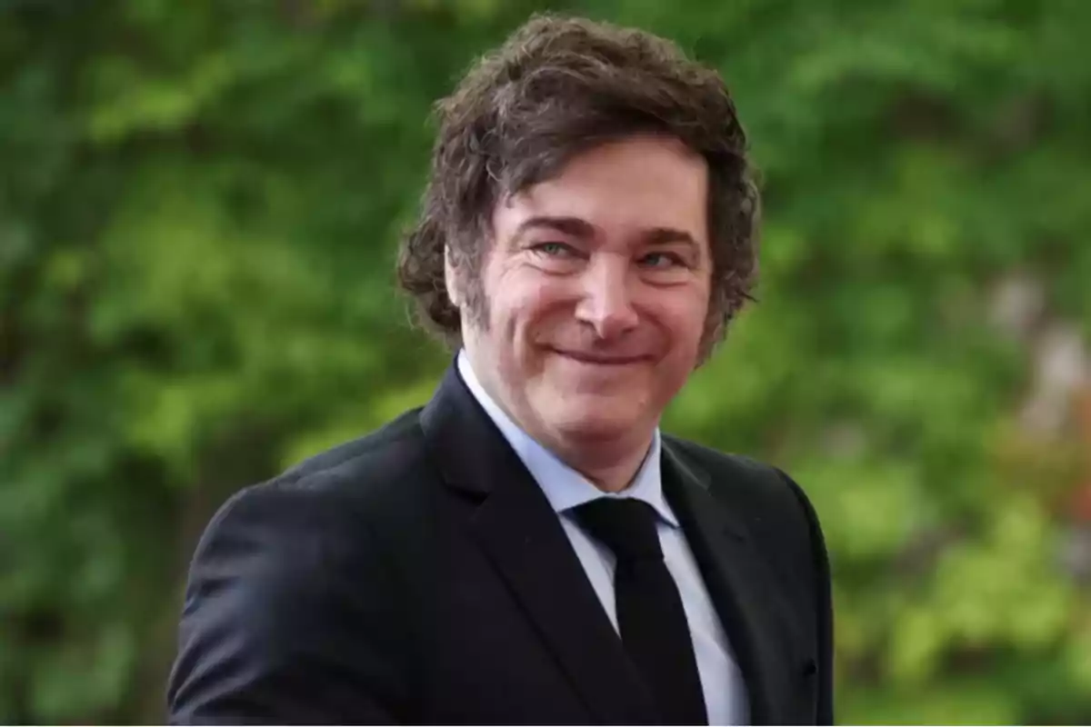 Hombre de cabello castaño y rizado vistiendo traje negro y corbata, sonriendo frente a un fondo de vegetación verde