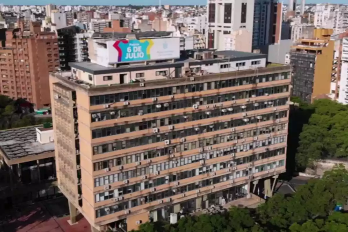 Edificio de varios pisos con numerosas ventanas y aires acondicionados en cada oficina rodeado de árboles y otros edificios en una ciudad