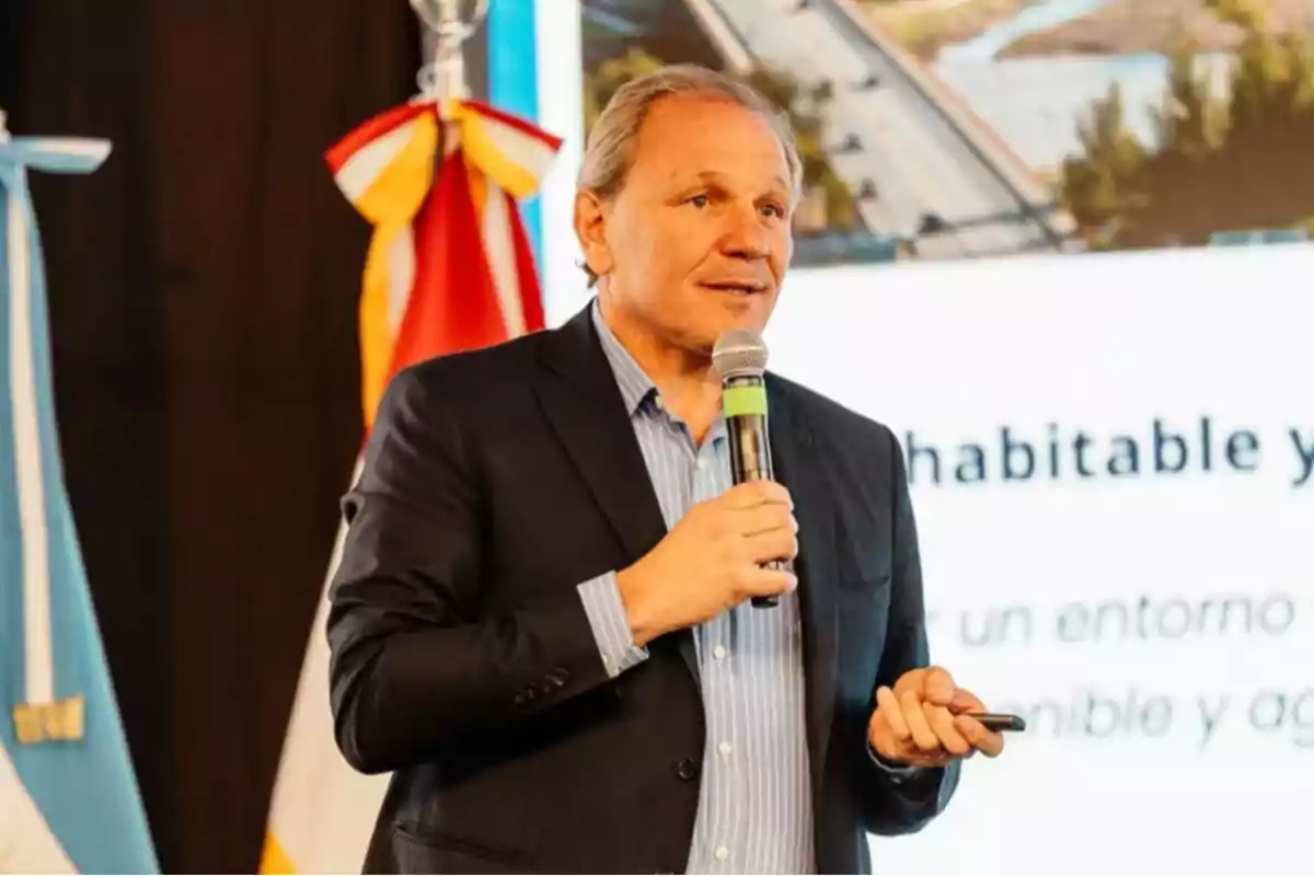Hombre de mediana edad con saco oscuro y camisa azul hablando en público con un micrófono en la mano y una pantalla de presentación de fondo