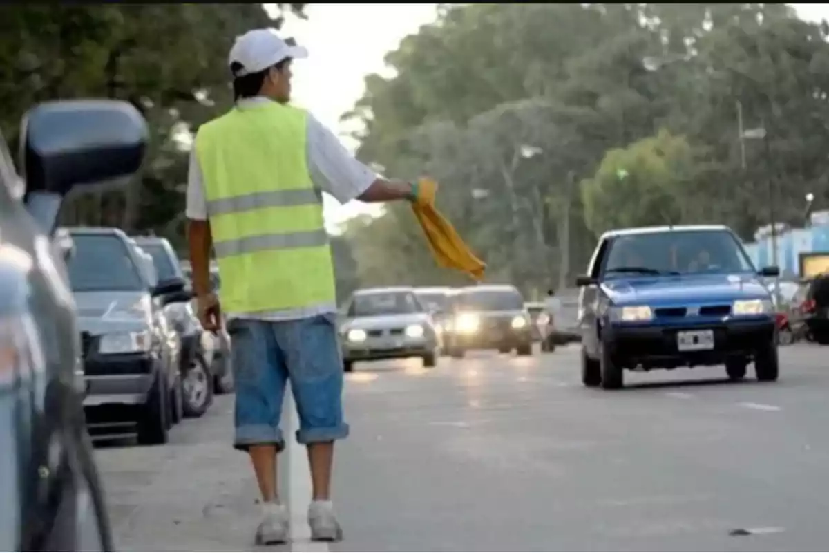 Persona con chaleco reflectante y gorra blanca guiando autos en una calle concurrida con varios vehículos y árboles al fondo