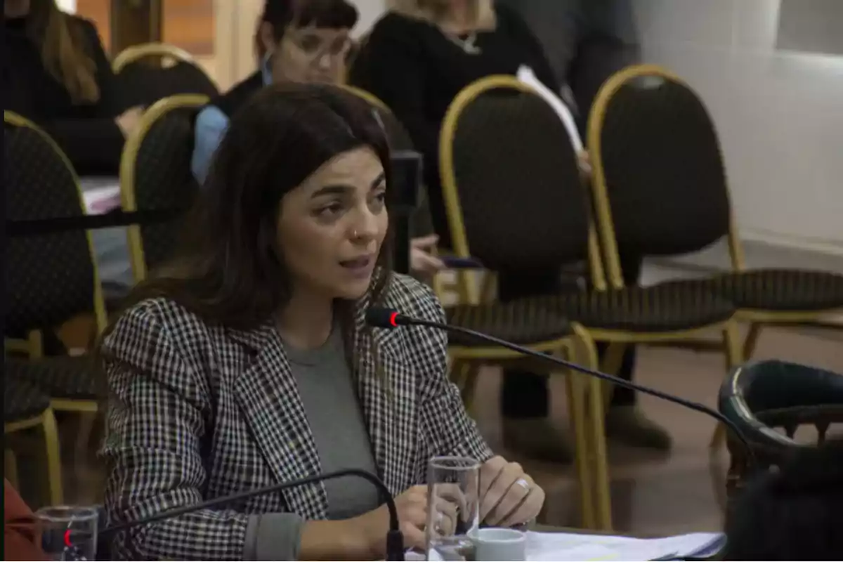 Mujer hablando frente a un micrófono en una sala con varias sillas y personas al fondo