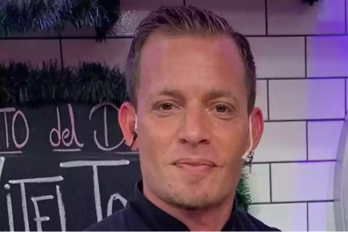 Hombre de cabello corto y rubio con auricular en la oreja, de pie frente a una pizarra en una cocina.