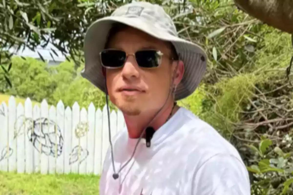 Hombre con sombrero gris y gafas de sol en un jardín con una cerca blanca al fondo Hombre con sombrero gris y gafas de sol en un jardín con una cerca blanca al fondo