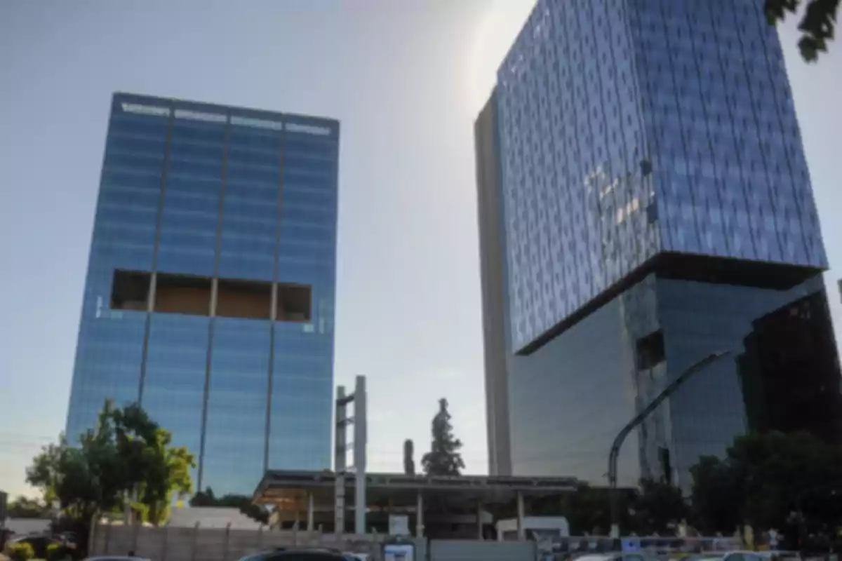 Dos edificios altos de cristal azul con árboles y una construcción en primer plano bajo un cielo despejado