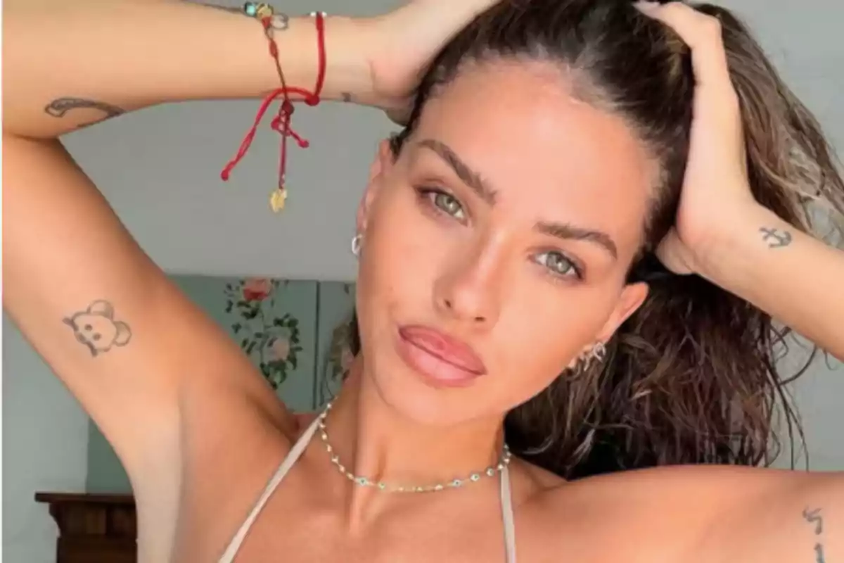 Mujer joven con cabello largo y mojado sostiene su cabeza con ambas manos mostrando tatuajes en los brazos y usando un collar delicado