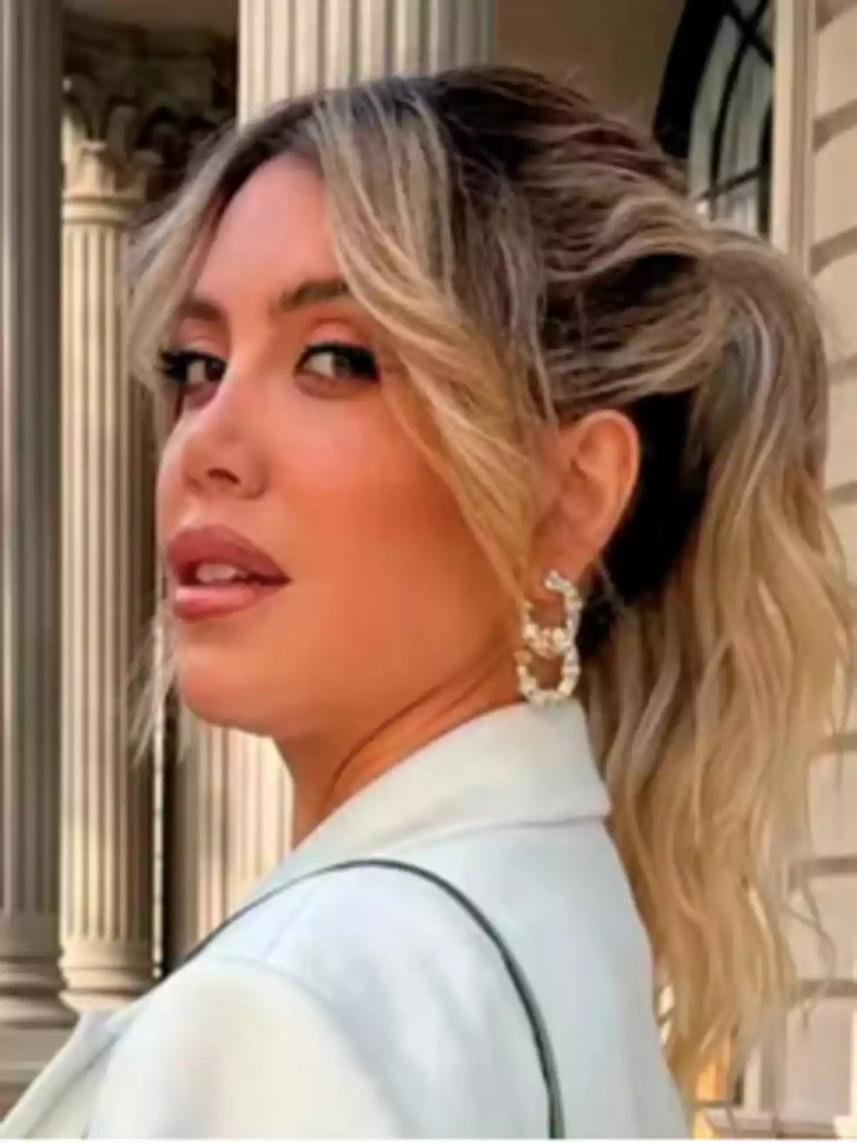 Mujer rubia con el cabello recogido y ondas suaves, usando un saco claro y aretes grandes, posando frente a un edificio con columnas. Mujer rubia con el cabello recogido y ondas suaves, usando un saco claro y aretes grandes, posando frente a un edificio con columnas.