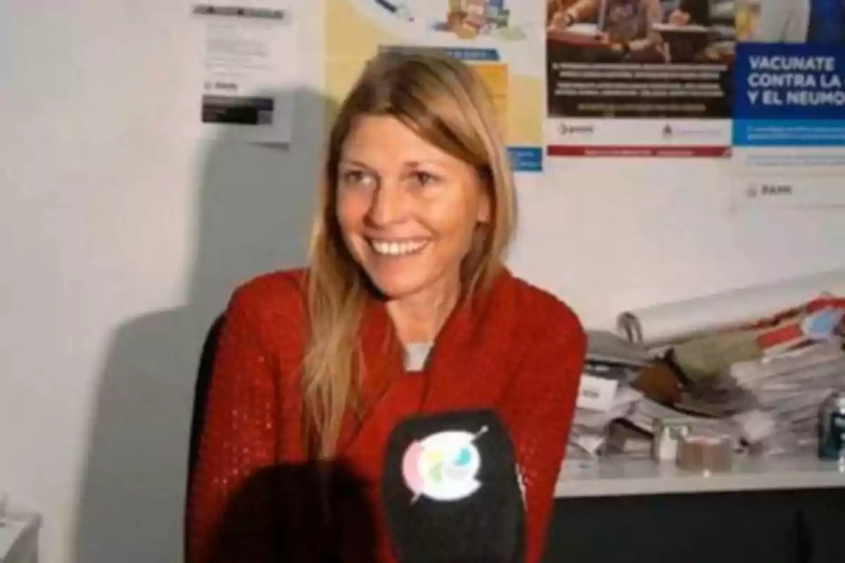 Mujer rubia con suéter rojo sonriendo frente a un micrófono en una oficina con papeles y carteles en la pared.