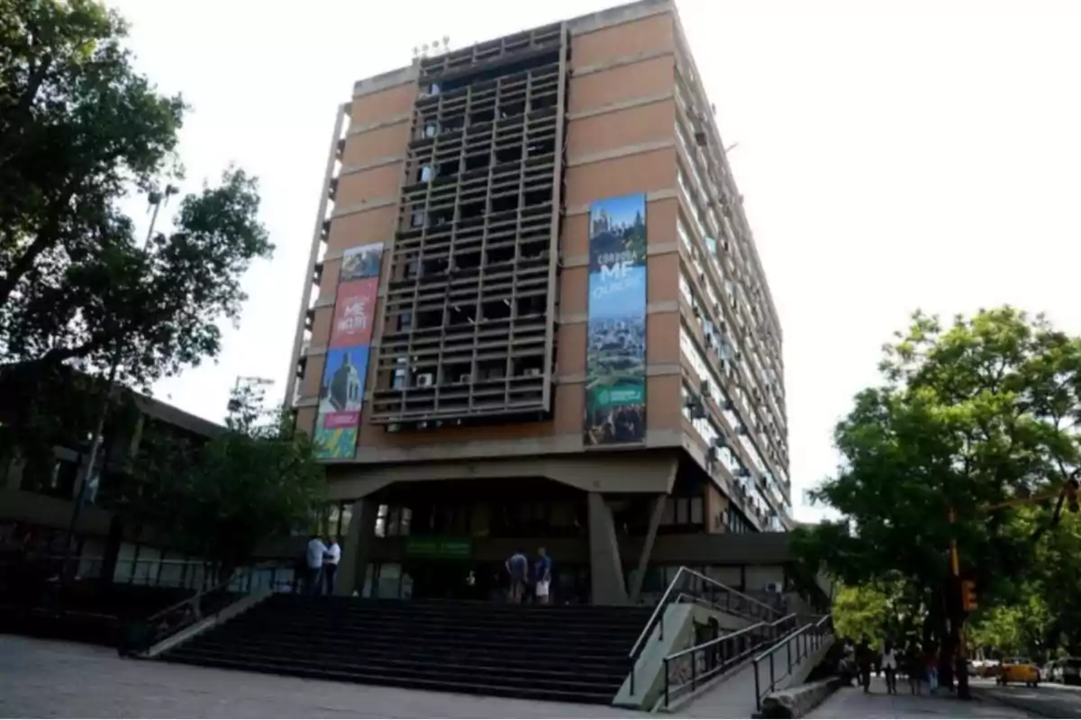 Multi-story building with a concrete structure and windows, surrounded by trees and with colorful signs on the facade, seen from the main entrance with stairs and people at the entrance. Multi-story building with a concrete structure and windows, surrounded by trees and with colorful signs on the facade, seen from the main entrance with stairs and people at the entrance.