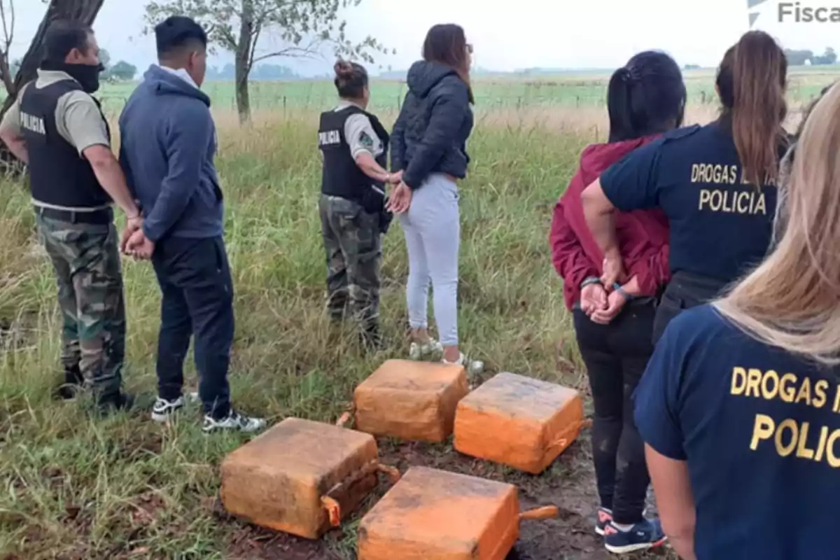 Varias personas esposadas son custodiadas por policías en un campo, junto a varios paquetes grandes envueltos en material naranja.