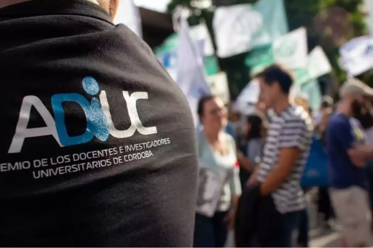 Person wearing an ADIUC t-jersey in the foreground during a protest, with several people and blurred flags in the background Person wearing an ADIUC t-jersey in the foreground during a protest, with several people and blurred flags in the background