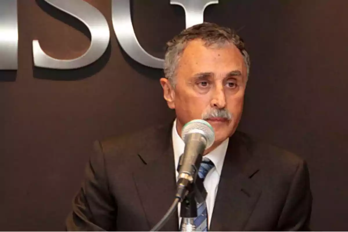 Hombre de cabello canoso y bigote hablando frente a un micrófono con fondo oscuro y letras plateadas detrás Hombre de cabello canoso y bigote hablando frente a un micrófono con fondo oscuro y letras plateadas detrás