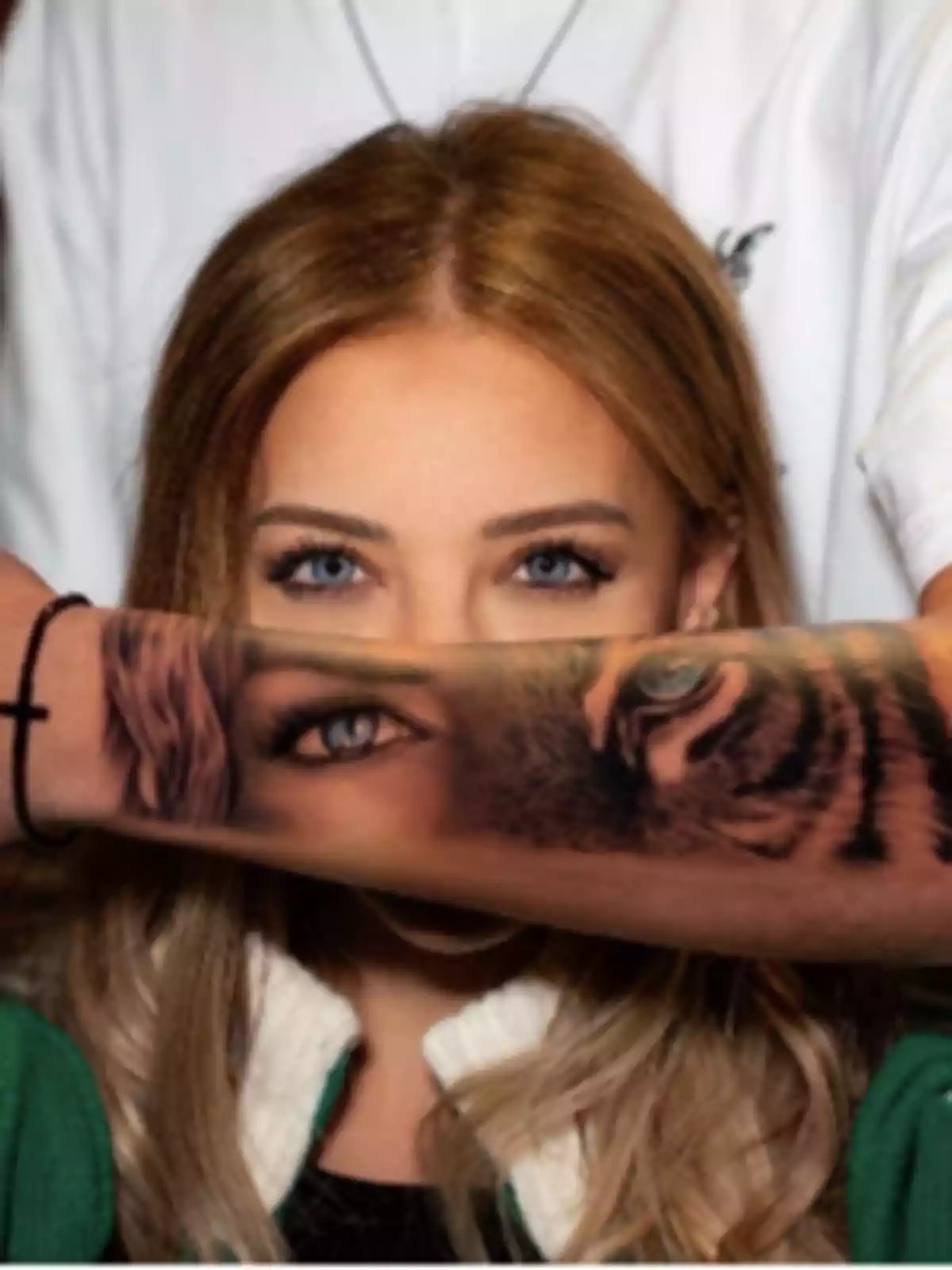 A light-eyed woman looks at the camera while a tattooed arm with an eye and a tiger’s face partially covers her face, aligning with her own eyes. A light-eyed woman looks at the camera while a tattooed arm with an eye and a tiger’s face partially covers her face, aligning with her own eyes.