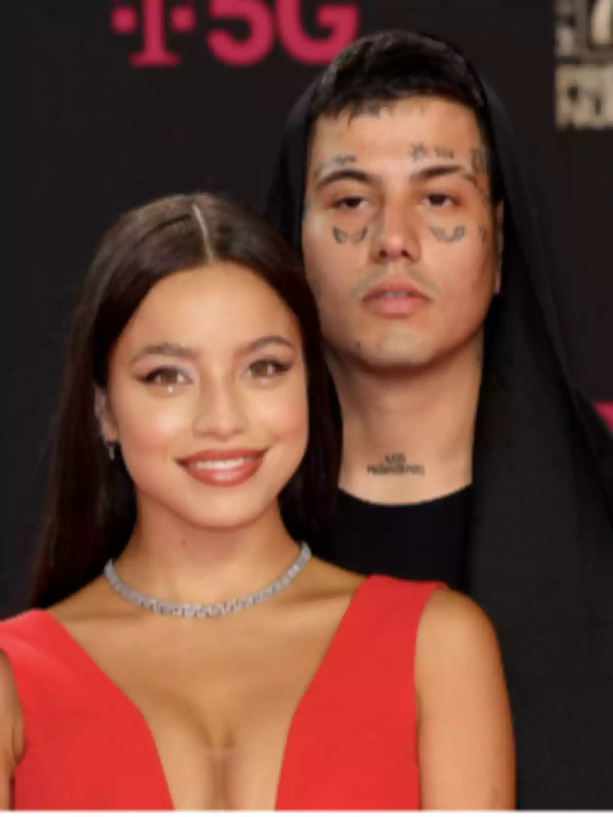 A woman in a red dress and silver necklace smiles in front of a man with facial tattoos and a black hoodie at a formal event. A woman in a red dress and silver necklace smiles in front of a man with facial tattoos and a black hoodie at a formal event.