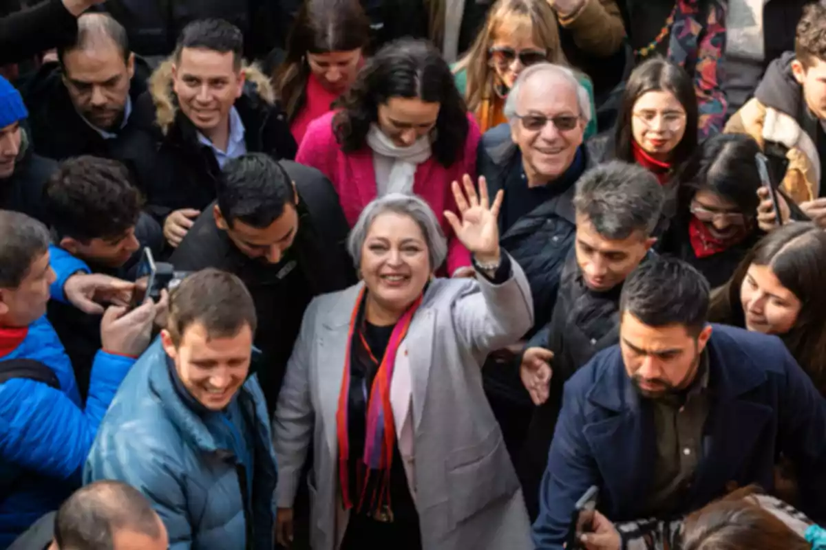 Mujer de cabello canoso sonriente saluda con la mano levantada mientras está rodeada de un grupo de personas en un ambiente al aire libre Mujer de cabello canoso sonriente saluda con la mano levantada mientras está rodeada de un grupo de personas en un ambiente al aire libre