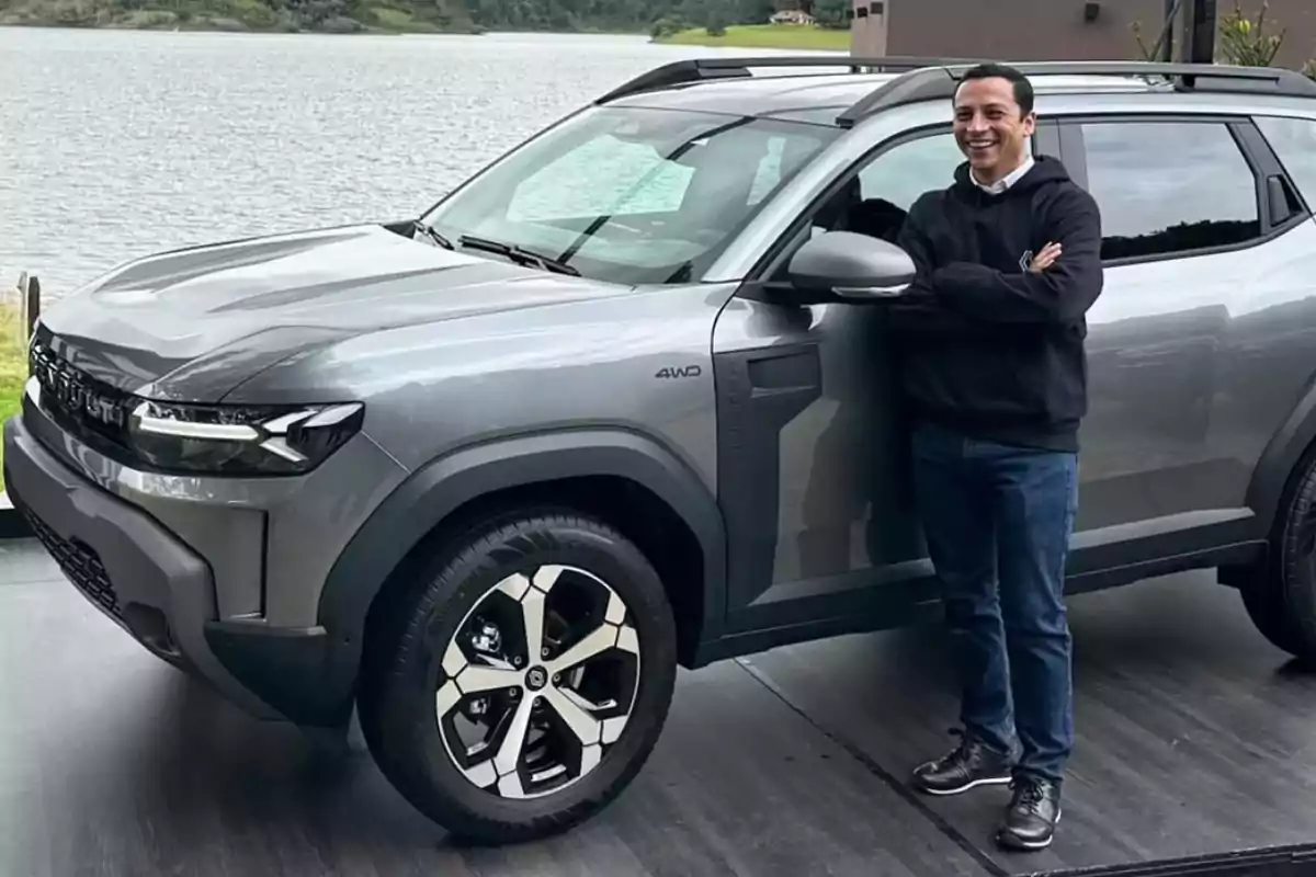 Hombre sonriente de pie junto a una camioneta gris estacionada cerca de un lago
